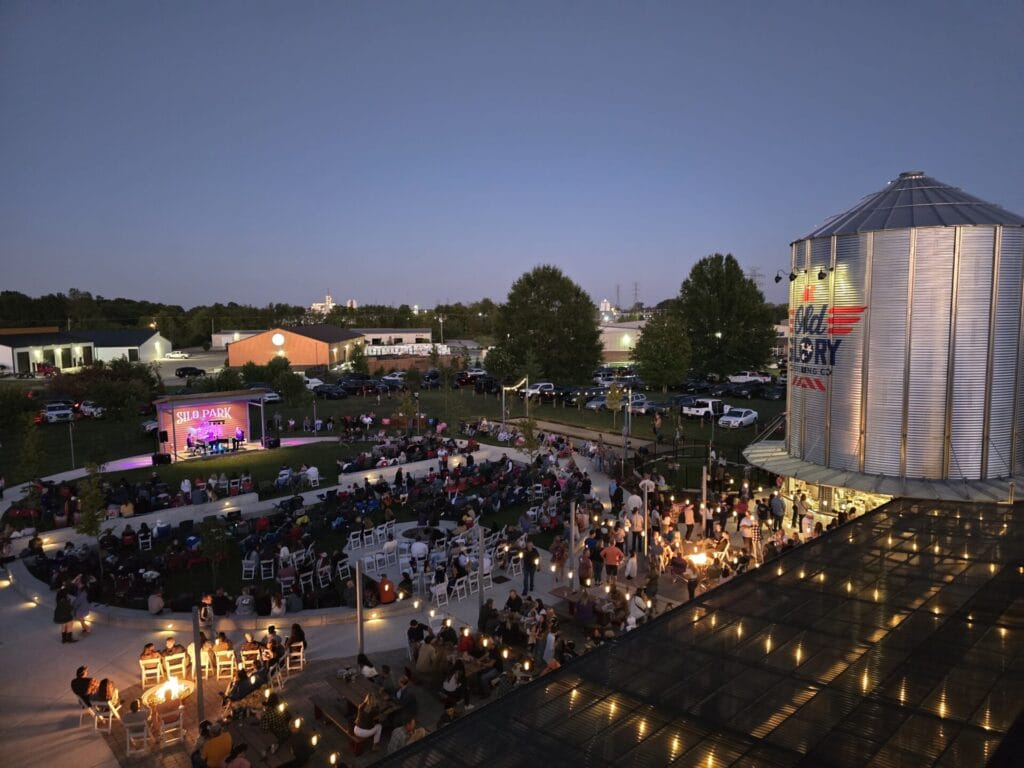 A lively evening at Silo Park at Old Glory Distilling Co. in Clarksville, TN, featuring an outdoor live music performance, guests enjoying firepits, craft cocktails, and a vibrant atmosphere under string lights.