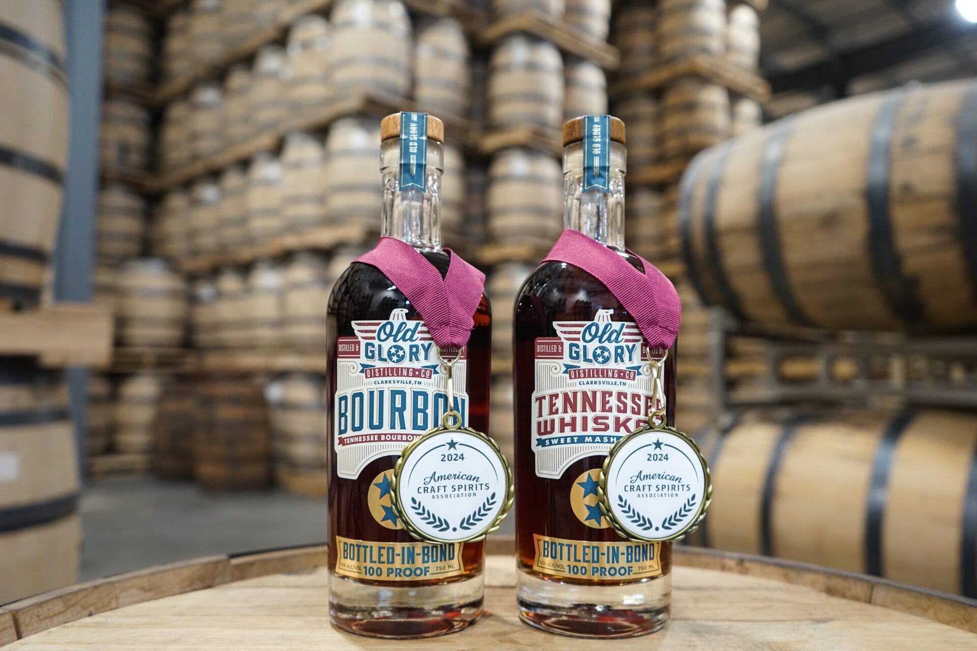 Two bottles of Old Glory Distilling Co.'s Bottled-in-Bond Tennessee Bourbon and Tennessee Whiskey, each adorned with a 2024 American Craft Spirits Association award medal. The bottles are set on a wooden barrel, with a background of stacked aging barrels in the distillery warehouse.