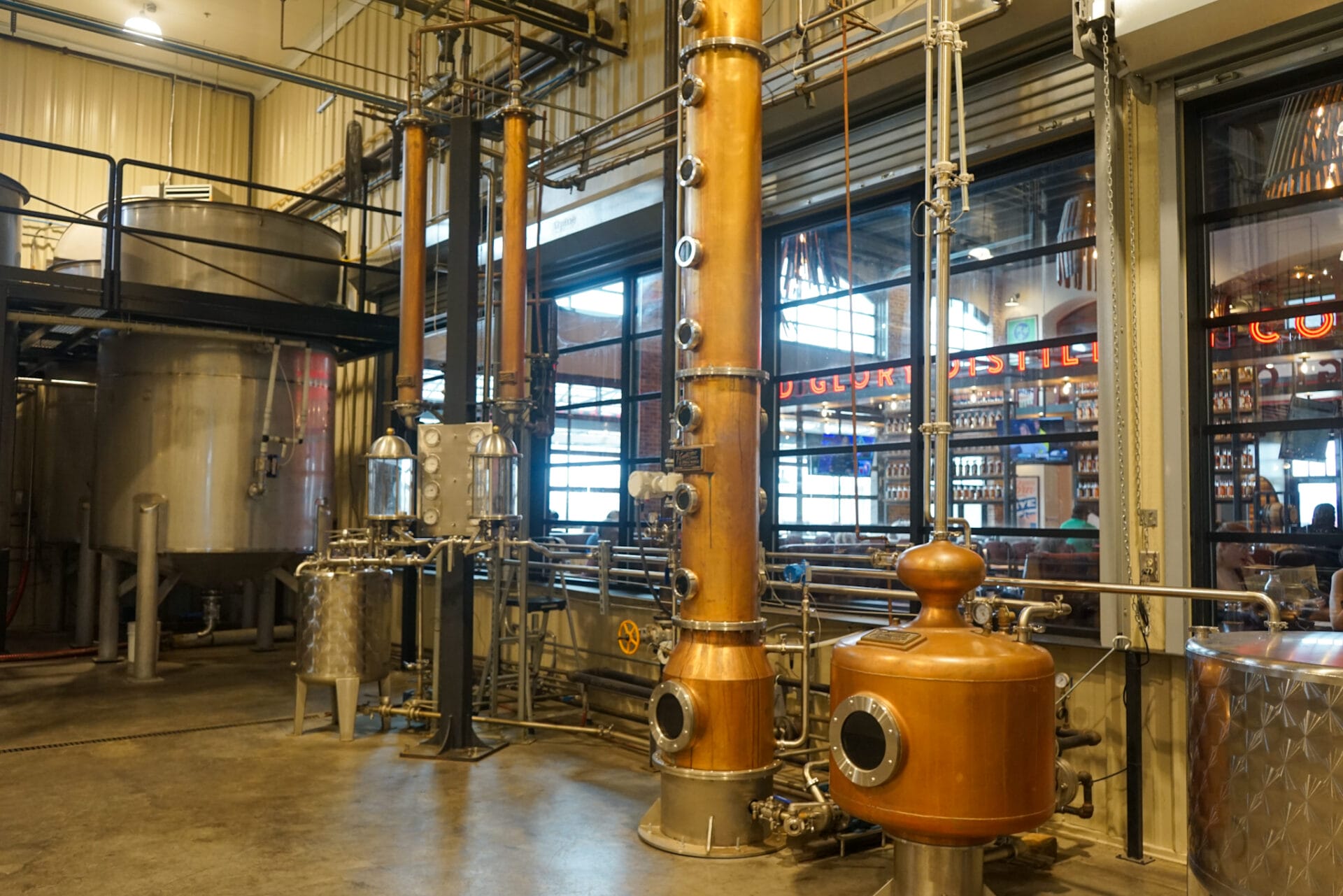 A copper column still and pot still inside Old Glory Distilling Co., used for whiskey production. Stainless steel fermentation tanks and additional distillation equipment are visible in the background, along with a windowed view of the distillery’s tasting room.