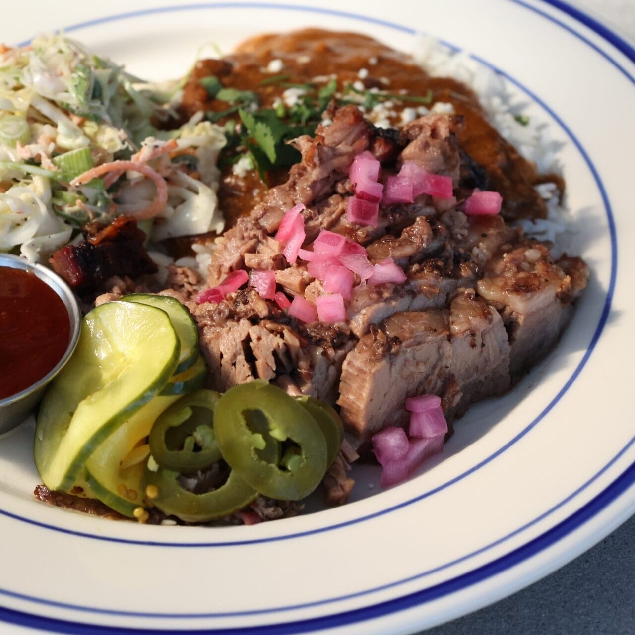 A plated barbecue dish featuring tender smoked brisket topped with pickled red onions, served with coleslaw, pickles, jalapeños, barbecue sauce, and a side of rice with rich sauce.